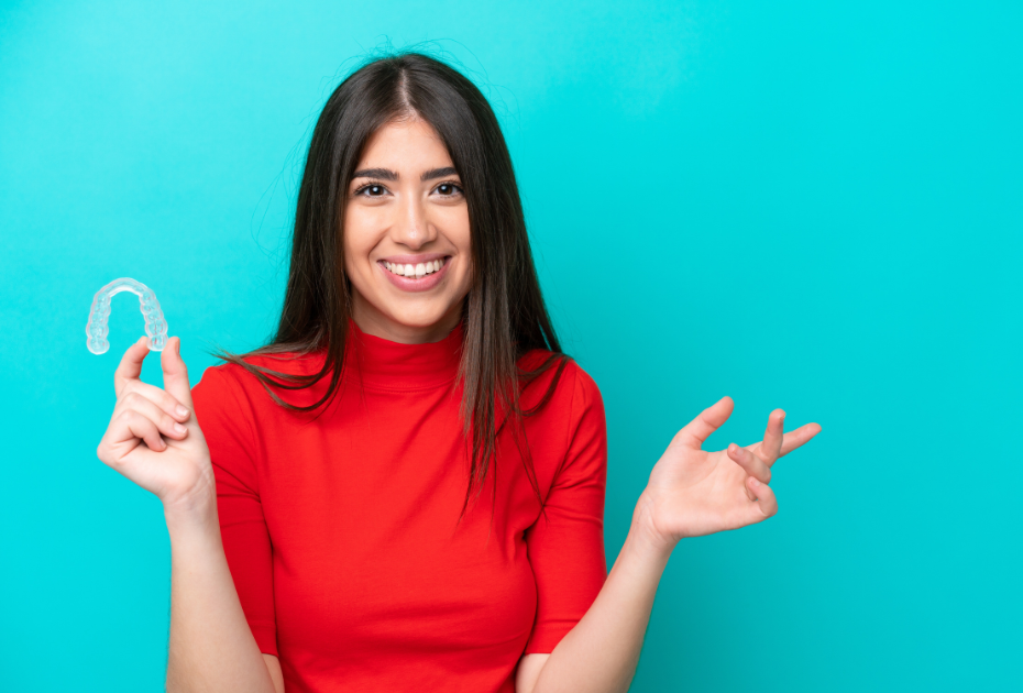 Smiling woman holding a clear dental aligner tray against a blue background, representing clear aligners vs braces in the UK.