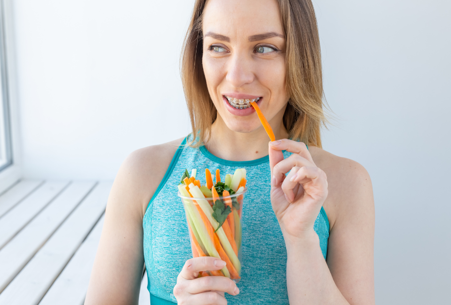 Woman eating carrot sticks while wearing clear aligners, holding a cup of fresh vegetables